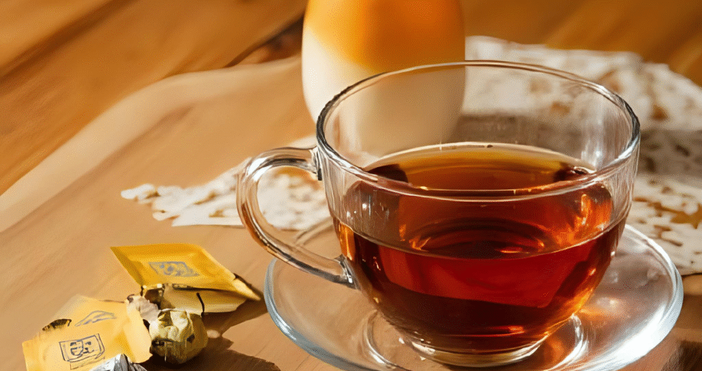 A glass cup of English breakfast tea on a saucer with tea bags and a milk jar on a wooden tray, representing a cozy tea setup.